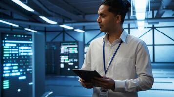 Programmer walking in high tech data center used for managing infrastructure energy consumption, working on tablet. Indian man using device in workspace used for automation in server farm, camera B photo