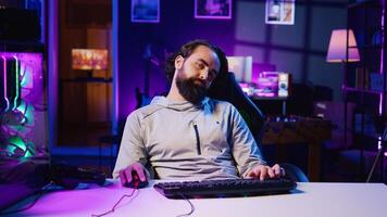 Drowsy man feeling tired after obsessively playing videogames all day, struggling to stay awake, unable to control addiction. Sedentary gamer falling asleep on chair while competing in online game photo