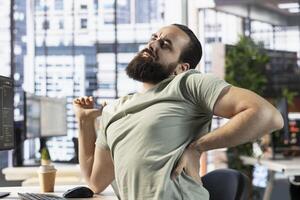 Tired employee stretching while checking key data needed for finding business insights and finishing project task. Fatigued man with sore back while analyzing data on computer photo