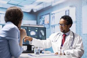 Female patient listening to a diagnostic report from doctor based on MRI scan results and x ray imaging, pointing at computer in medical cabinet. African american expert gives assistance. photo