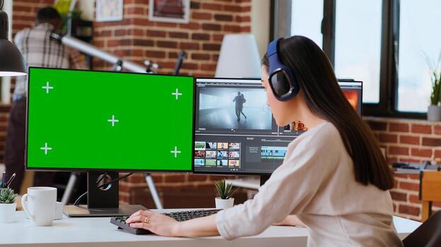 Professional asian woman working on a media project next to green screen in a modern space, using dual monitors and advanced editing software. Multitasking in post production. Camera A. photo