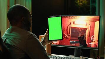 Man watching videos on green screen tablet while playing first person shooter videogame at home. Gamer looking at chroma key device display while enjoying online game on PC photo