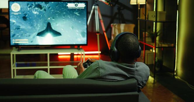 Man playing entertaining spaceship flying videogame on large smart TV, wearing headphones. Player enjoying SF arcade game on gaming console, piloting spacecraft in galaxy using gamepad, camera B photo
