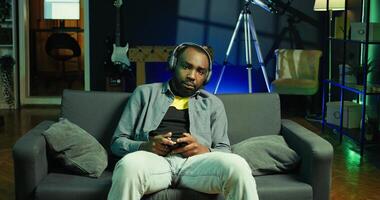 Tired gamer in RGB lit home late at night gaming with gamepad. Man sitting on sofa in neon illuminated apartment, playing videogames on console using controller, looking exhausted, camera B photo