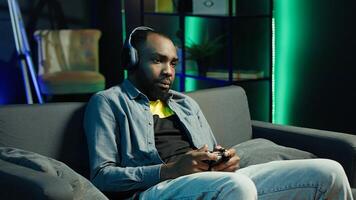 Tired gamer in RGB lit home late at night gaming with gamepad. Man sitting on sofa in neon illuminated apartment, playing videogames on console using controller, looking exhausted, camera A photo