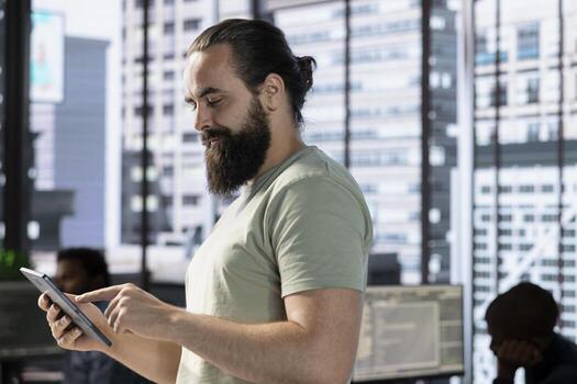 Startup employee walking in office, using tablet to do research for project with deadline approaching. Man using digital device in workspace, solving tasks during job shift, city backdrop photo