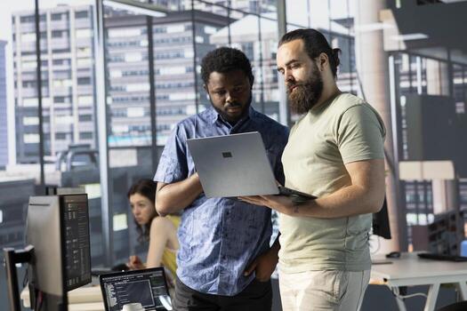 IT specialists in office working together using laptop, testing and deploying programs and systems. Programmer and coworker checking code on notebook, doing brainstorming in workspace photo
