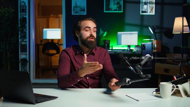 Content creator talking in front on cellphone attached to selfie stick in studio, chatting with viewers using recording mic. Man using phone camera and professional microphone to record vlog, camera B photo