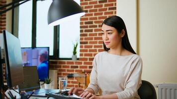 IT specialist types code on dual monitors in a cozy home office while working remotely. Asian woman uses programming language for productivity in coding and internet security. Camera A. photo