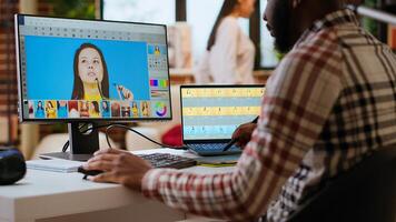 African american male designer edits images on dual monitors, computer setup in a creative office. Ideal workspace for digital media professional focused on photo retouching. Camera B.
