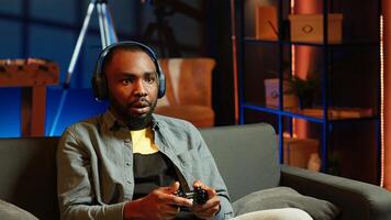 Cheerful man wearing headphones, celebrating after victory in videogame, neon lit apartment. Person on couch throws fists in air from excitement after winning game on console with controller, camera A photo