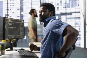 Worker stretching to relieve muscle strain while conducting code review to fix bugs, identifying technical issues with software. Man in office suffering back pain from bad posture, optimizing code photo