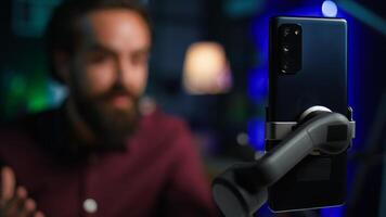 Focus on cellphone used by media star using on selfie stick to film vlog channel intro. Influencer greets viewers, presenting today topic, recording with mobile phone, camera B close up photo
