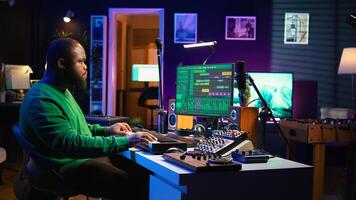 Sound engineer pressing keys of electronic synthesizer and composing new song, adjusting volume levels on pre amp knobs. Composer producing music on piano midi controller, mixing tunes. Camera B. photo