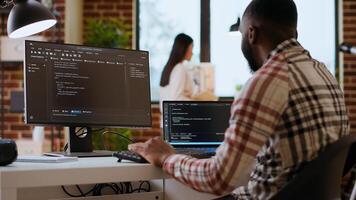 African american IT professional types programming code on dual monitors in home office, working on software development and internet security. Ideal setup for remote work and productivity. Camera B. photo