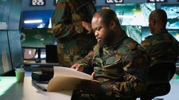 Military soldier in mission control center overseeing national safety operations, reading folder files. African american army specialist checking data in monitoring room command center, camera A photo