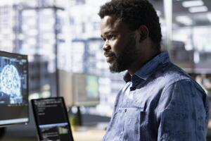 Cheerful employee utilizing AI cloud computing resources and platforms to train and deploy large scale machine learning models. Smiling african amertican man monitoring and maintaining company systems photo