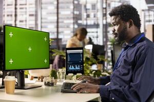Black businessman at office desk works on daily tasks next to green screen on device, studying analytics and infographics to support effective decision making for success. Business operations. photo