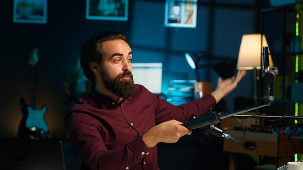 Influencer doing peace sign in front on cellphone attached to selfie stick in studio, chatting with viewers using mic. Man doing hand gesturing, using phone and microphone to record vlog, camera A photo