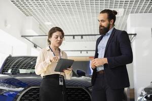 Car dealership saleswoman asking man questions to determine make, type and quality of vehicle desired. Employee ensuring clients satisfaction, answering inquiries, showing auto model on tablet photo