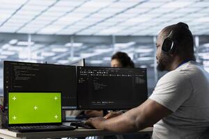 Isolated screen laptop in front of IT support workers coding on computers and listening music. Engineer wearing headphones at PC desk, overseeing server clusters using chroma key notebook photo