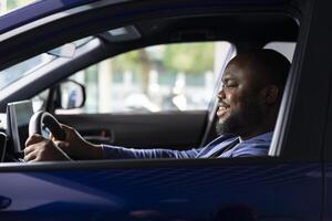 Smiling man enjoying car test drive in showroom, making sure he feels comfortable and enjoys driving vehicle. Cheerful african american client trying out new automobile before making investment, photo
