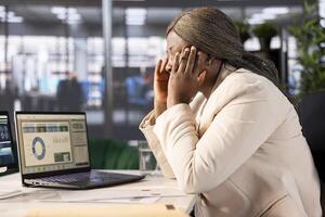 Overworked woman having migraine symptoms while looking over statistics charts on laptop, researching data. Tired worker in pain, feeling discomfort while doing research for marketing project photo