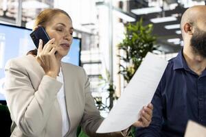 Woman in workspace talking on phone and looking at financial business documents. Worker in office checking accounting figures, finishing tedious paperwork, having telephone call photo