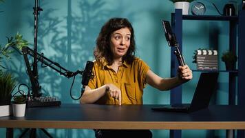 Smiling woman in studio using smartphone on selfie stick to record , greeting subscribers. Joyous entertainer hosts q and a session with viewers using cellphone, saluting fans, camera B photo