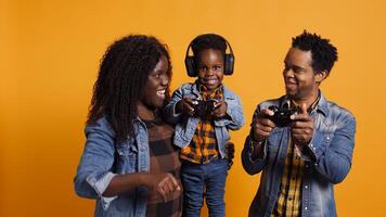 Enthusiastic father playing online games with baby boy using controller, both parents enjoying leisure time with their small kid. African american happy family play with joystick. Camera B. photo