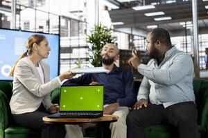 Multiethnic colleague using green screen laptop, doing briefing meeting to share new ideas for profit growth. Diverse team of employees in office talking with director, using chroma key notebook photo