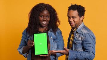 African american couple pointing at a tablet with isolated green screen display, smiling at each other against yellow background. Cheerful affectionate husband and wife posing. Camera A. photo