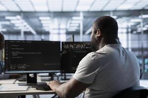 African american admin programming in server room housing advanced equipment storing massive datasets. Engineer typing code on PC to mend data center racks doing complex computational operations photo