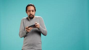 Focused man plays intense videogame on mobile phone, spamming attack buttons on touchscreen. Gamer participates in gaming competition, isolated over studio background, camera B photo