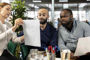 Multiethnic colleague doing briefing meeting to share new ideas for profit growth, presenting documents. Diverse team of employees in office talking with director, showing progress done on project photo