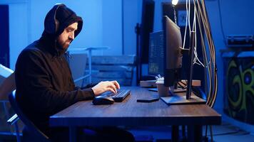 Hacker listens music while hiding in underground bunker and coding, stealing data from victims using PC virus. Rogue programmer wearing headphones tricks people into installing ransomware, camera A photo