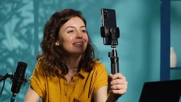 Smiling woman in studio using smartphone on selfie stick to record , greeting subscribers. Joyous entertainer hosts q and a session with viewers using cellphone, saluting fans, camera A photo