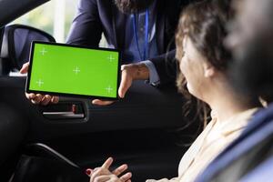 Car salesman showing product offerings on green screen tablet to woman, making recommendations. Showroom agent presents financing options to customer during test drive using chroma key device photo