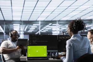 Team of programmers in server farm using green screen laptop, looking for misconfigurations. African american IT experts mending data center bugs with chroma key notebook photo