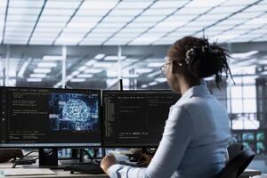 Engineer working at PC desk, doing checkup on AI server room providing computing resources. African american IT professional investigating data center mainframes using computer photo