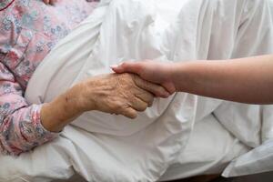 A young womans hands gently hold the hands of an elderly female patient in a hospital bed, symbolizing care, support, and compassion. photo