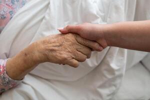A young womans hands gently hold the hands of an elderly female patient in a hospital bed, symbolizing care, support, and compassion. photo