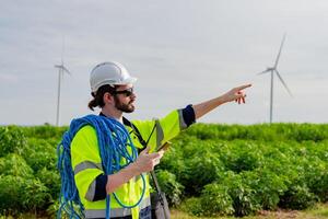 Worker in reflective gear points at wind turbines in a green field while holding equipment and ropes photo