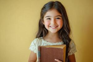 portrait of a cute smiling girl holding book on yellow background with copy space photo