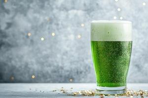close-up glass of green beer with foam on a light gray concrete background with subtle golden confetti with copy space photo
