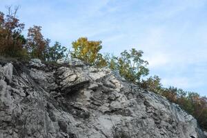 Rocky cliff with patches of green and autumn trees under a partly cloudy blue sky depicting natural erosion and geology. photo