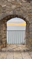A rustic stone archway frames a view of calm blue waters and a golden sunset, with a black iron railing in the foreground. High quality photo