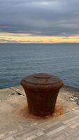 A large rusted mooring bollard on a stone pier with calm sea waters and a dramatic sunset sky in the background. photo