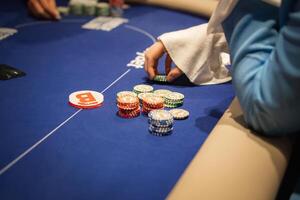 A poker game in progress on a blue felt table with multicolored chips, a dealer button, and a player's hand reaching toward a chip stack. photo