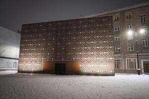A winter evening in Riga, Latvia, with a snow covered square, a patterned building with geometric facade, and a lit streetlamp near a brick structure. photo